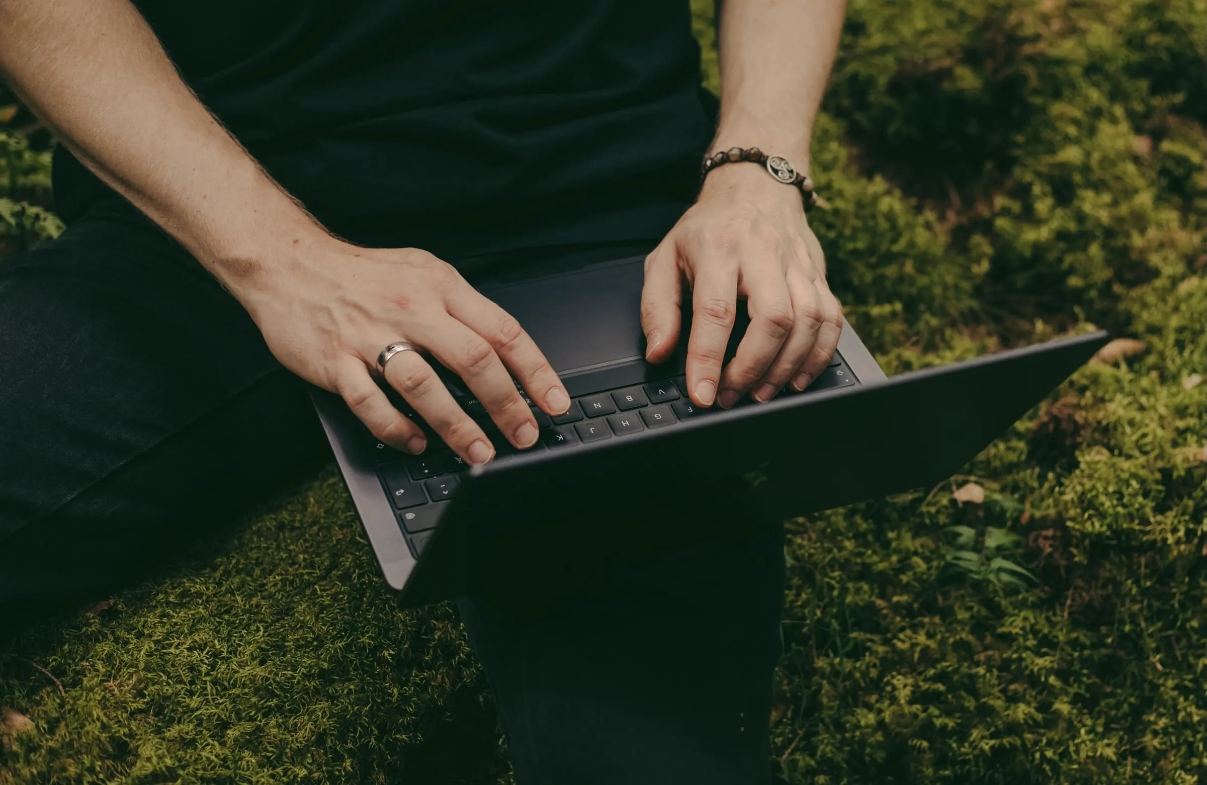 A person typing on a laptop outdoors on mossy ground